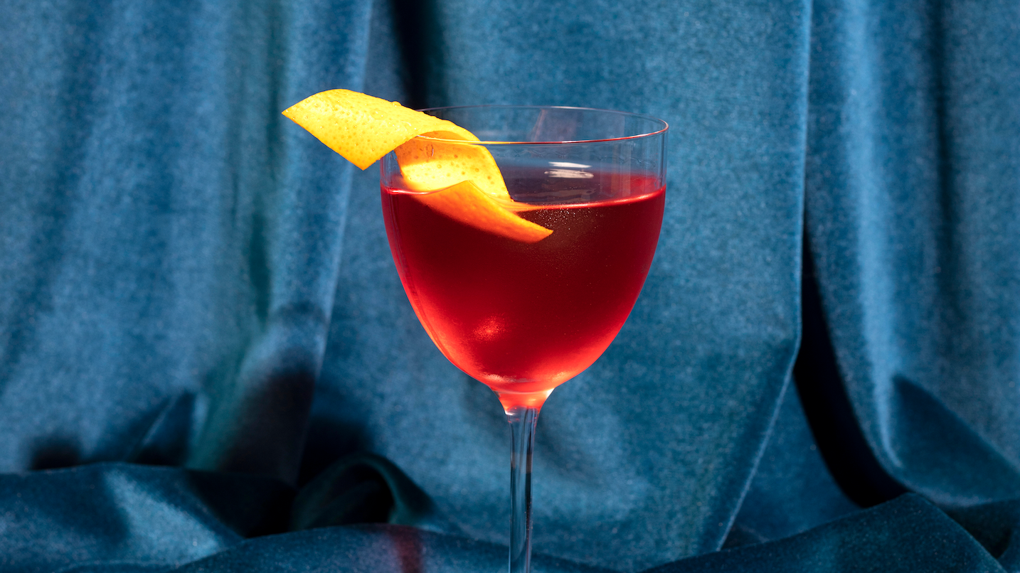 A red cocktail in a coupe glass sitting on a blue velvet curtain backdrop. A yellow lemon peel is twisted ontop for garnish.