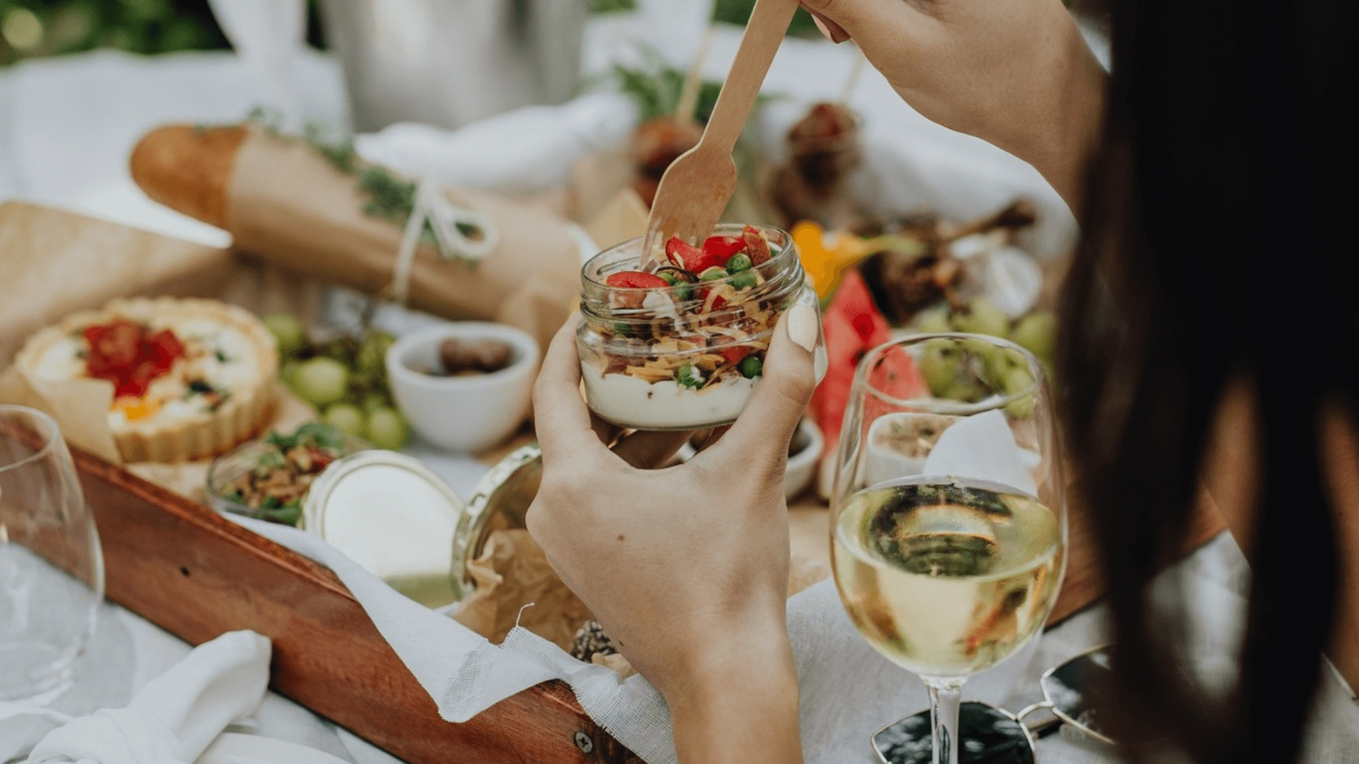 A women sits at a picnic table, eating a selection of dishes with a glass of wine.