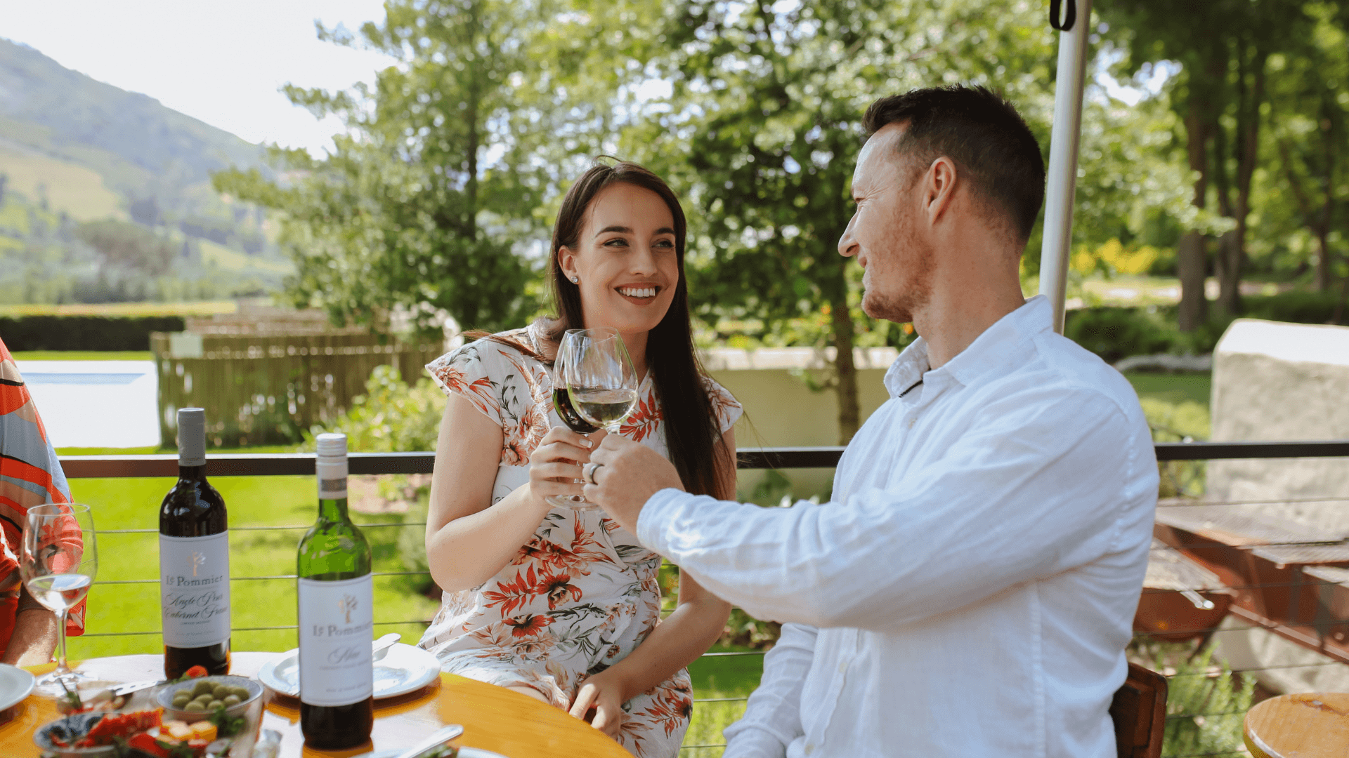Valentine’s Day specials in South Africa 2026 Two people cheers with a glasses of wine. A mountain and trees are in the background.