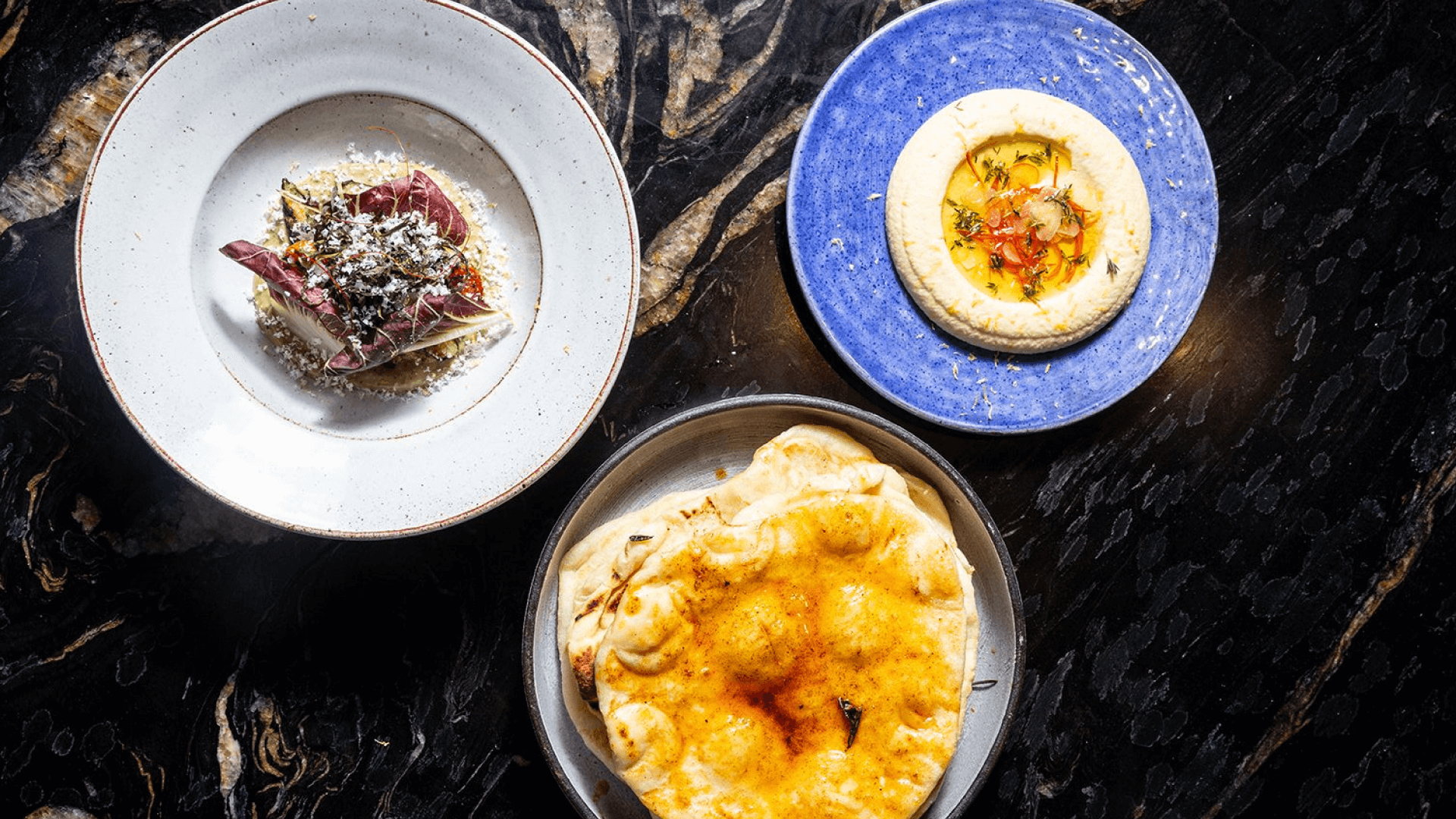Flat lay of three plated dishes on a dark marble surface: hummus topped with oil and herbs on a blue plate, charred flatbread with spices in a metal bowl, and a vegetable salad with radicchio, grains, and grated cheese on a white plate.