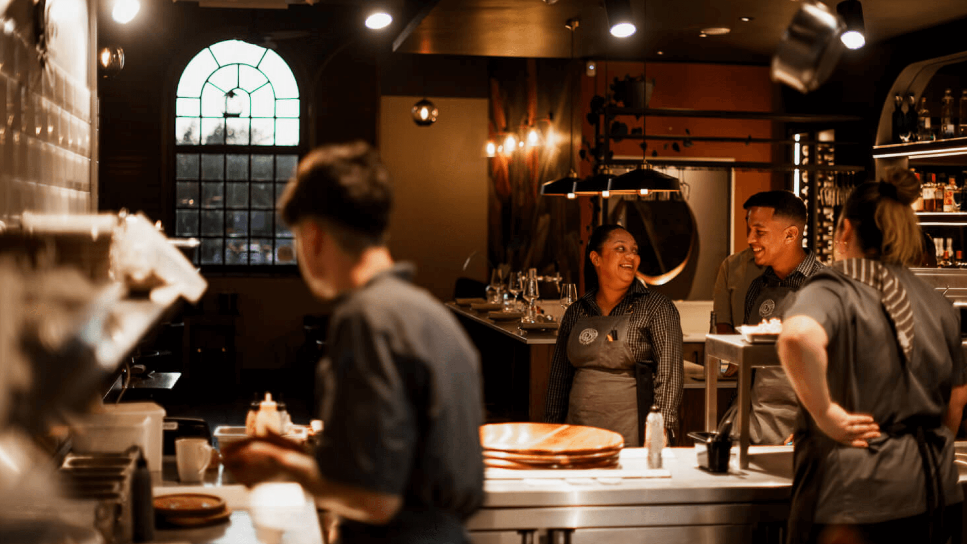 Restaurant staff in uniform chat and smile in a warmly lit kitchen.