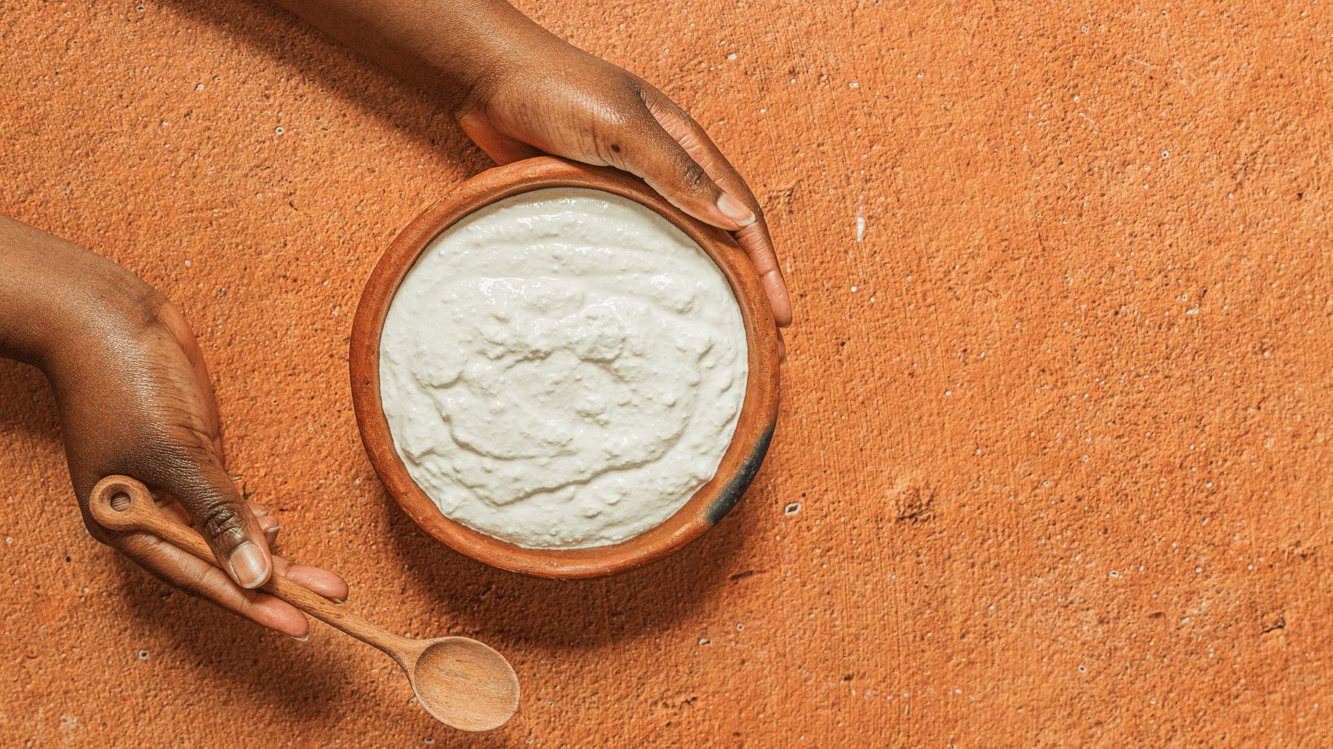 A woman holds in her hands a bowl of amasi and a wooden spoon on a orange clay floor.