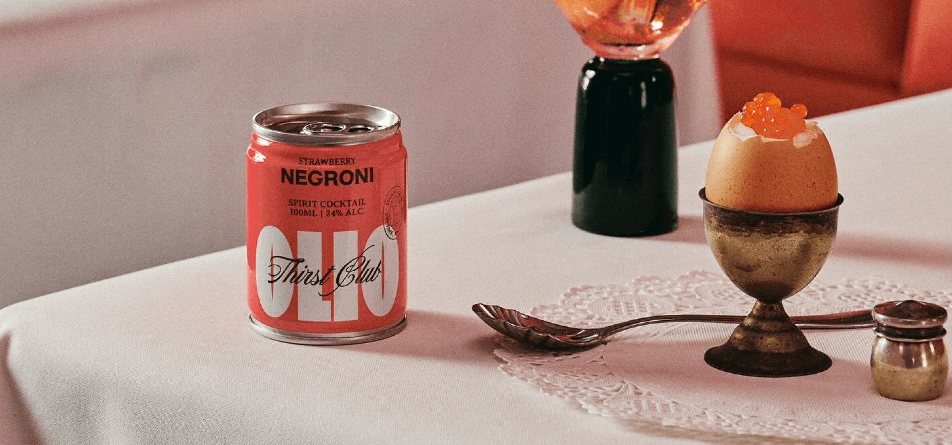 A small can of Olio Cocktail Negroni sits on a white table next to a hard-boiled egg in a silver egg holder. The egg is topped with orange caviar. 