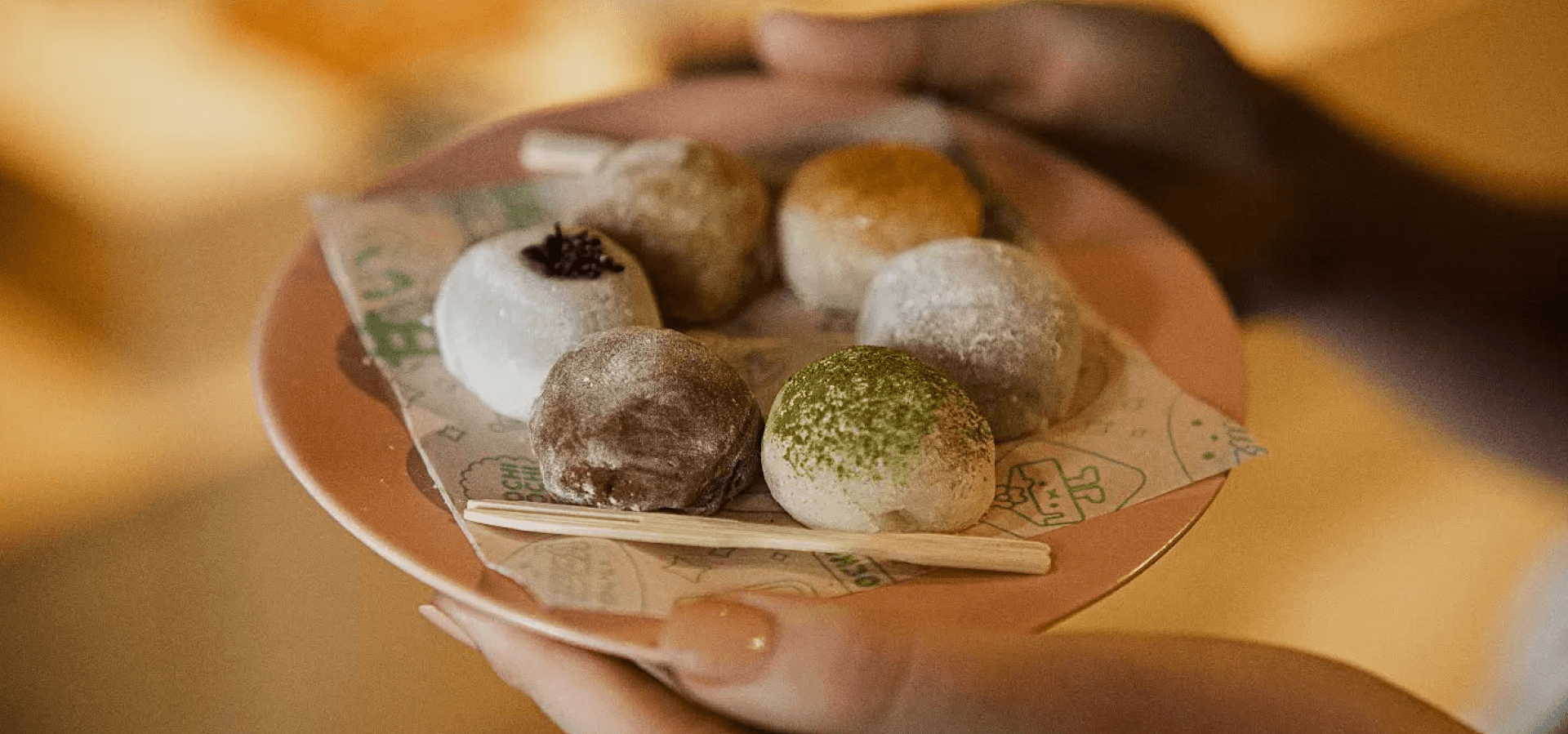 A woman holds a pink plate that has 6 different colours of mochi on it.