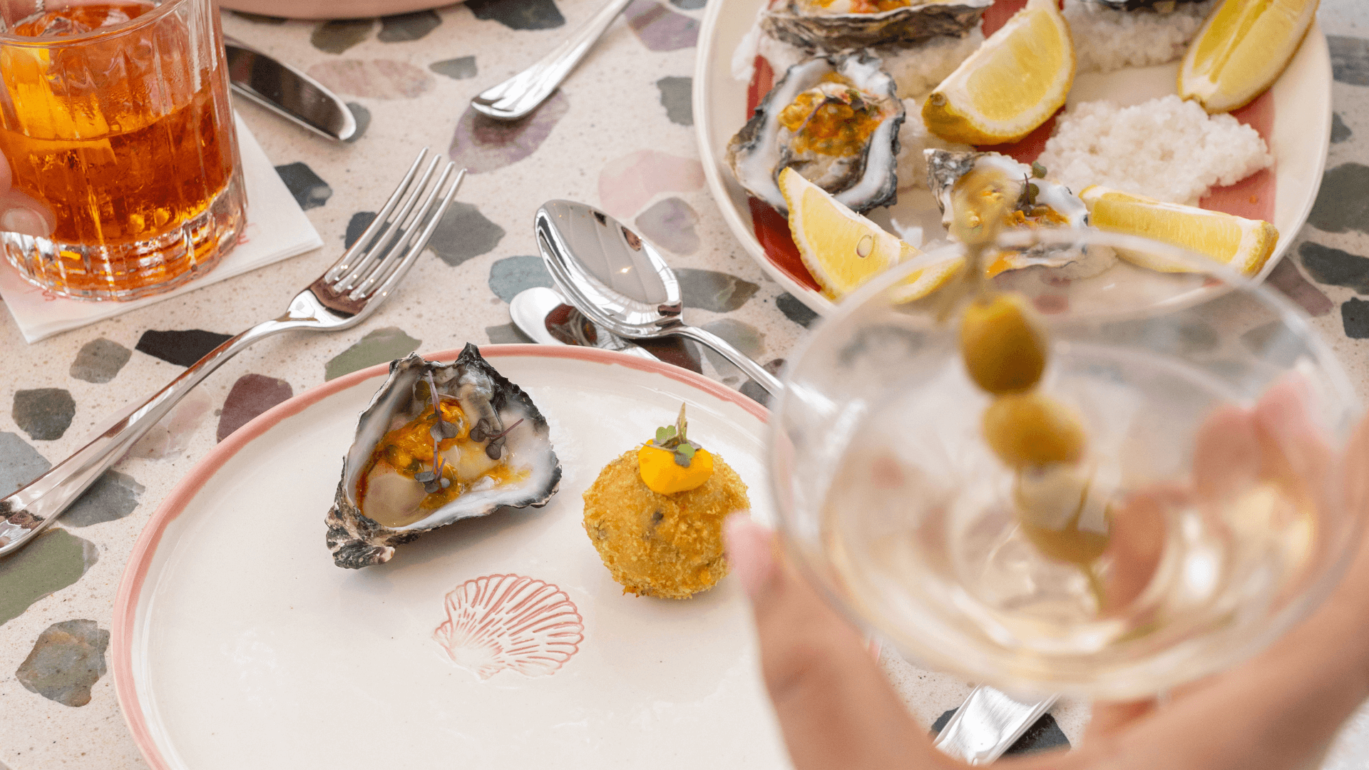 A martini with olives is blurred in the forefront, behind, oysters and an orange cocktail are on a table and pink plate.